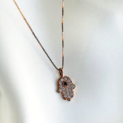 An 18 Karat rose gold Hamsa pendant with pave-set round brilliant cut diamonds and a bezel-set blue sapphire center, attached to a 14 Karat rose gold chain.