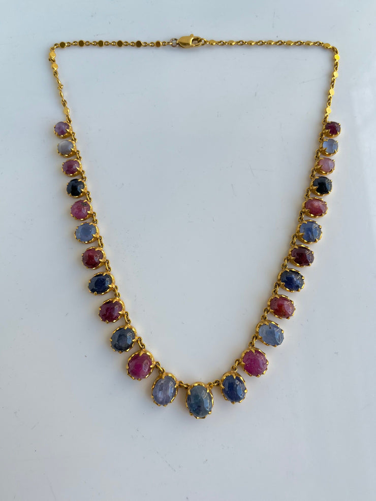 A complete, laid-flat image of the necklace captures its graceful choker-length design, highlighting the rhythmic pattern of alternating sapphire and ruby cabochons. The warm glow of 18K yellow gold provides a rich backdrop for the vivid gemstones, creating a regal and balanced aesthetic.