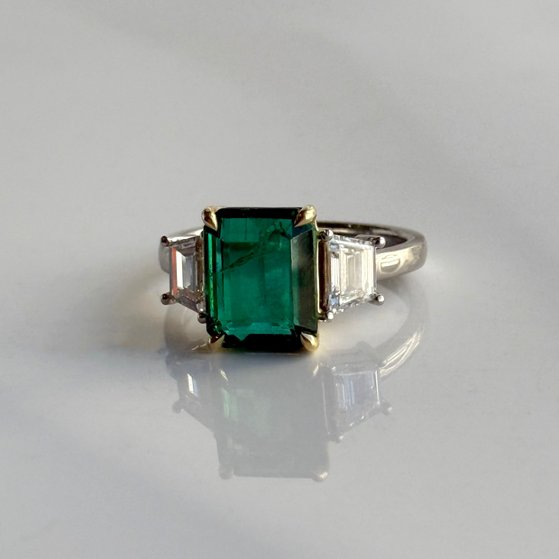 Ring with a green emerald gemstone and trapezoid diamond side stones on a white background