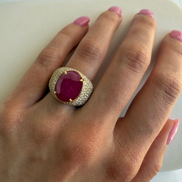 Hand wearing a ring with a red ruby gemstone and gold diamond band on a white background