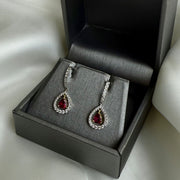 Teardrop-shaped diamond earrings with red ruby gemstones in a gray jewelry box on a light background