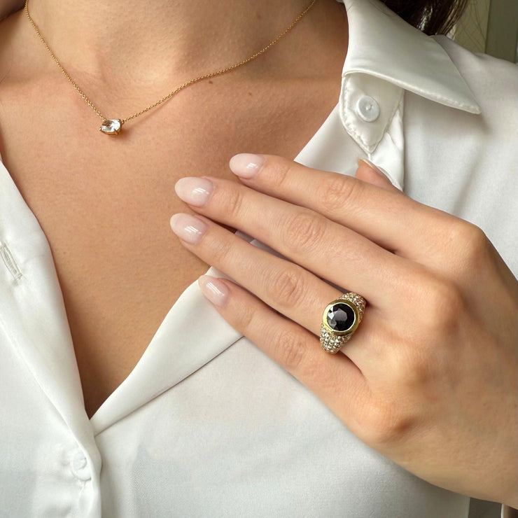 Close-up of a hand wearing a gold sapphire dome ring, with a diamond necklace and white silk shirt in the background.