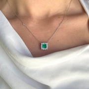 A model elegantly wearing an 18k white gold necklace featuring a 1.51 ct light green emerald surrounded by a diamond halo. The necklace catches the light, enhancing the emerald's vibrant hue and the sparkle of the diamonds, adding a touch of sophistication to the model's outfit.