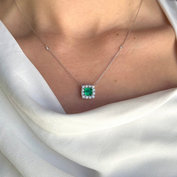 A model elegantly wearing an 18k white gold necklace featuring a 1.51 ct light green emerald surrounded by a diamond halo. The necklace catches the light, enhancing the emerald's vibrant hue and the sparkle of the diamonds, adding a touch of sophistication to the model's outfit.