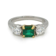 Emerald at the center set in yellow gold, flanked by two round brilliant cut side diamonds set in white gold.