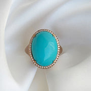 Front view of a turquoise cabochon ring with diamond halo in matte 18k rose gold against silk