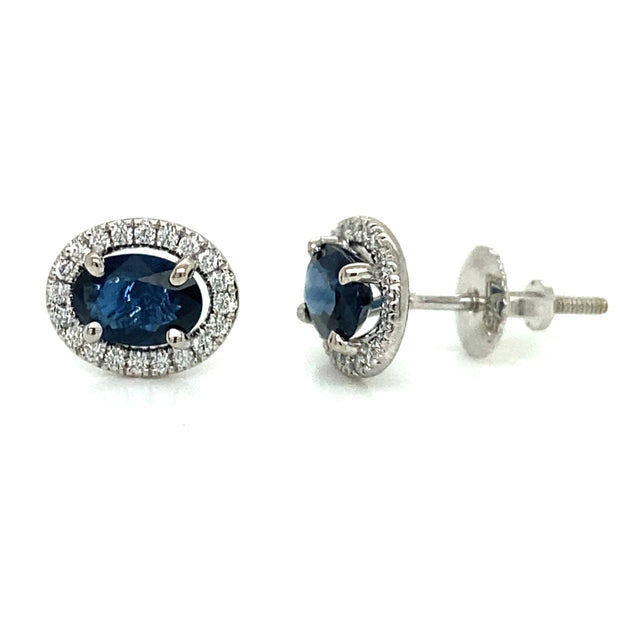 A pair of 18K white gold stud earrings, featuring vibrant oval sapphires surrounded by diamond halos.