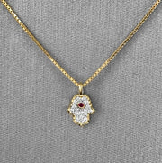 An 18 Karat Yellow Gold petite Hamsa pendant with pave-set round brilliant cut diamonds and a bezel-set ruby at the center, attached to a 14 Karat Yellow Gold chain.